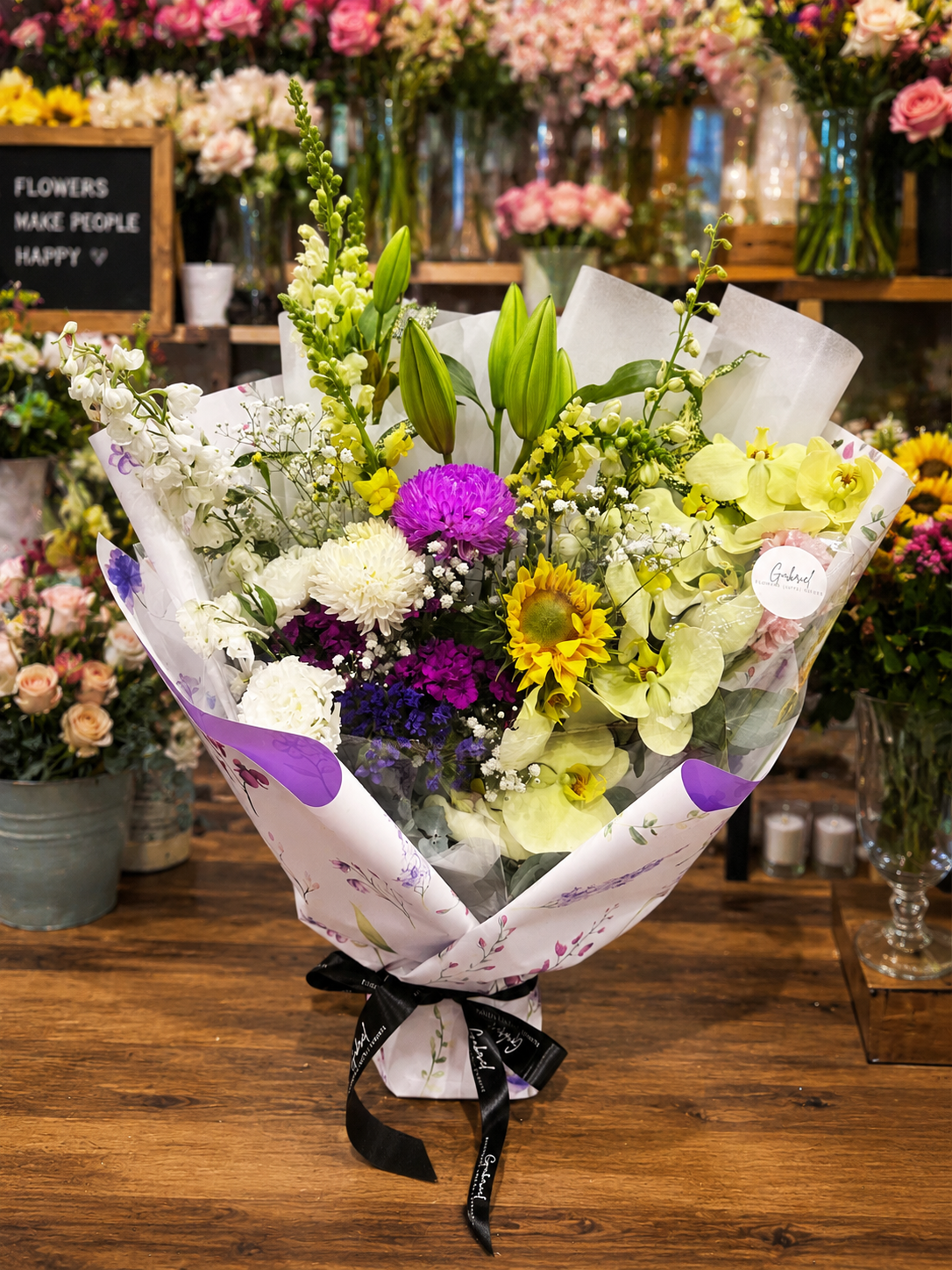 Bouquet of flowers with orchid, lilies, sunflowers, chrysanthemum, with a black ribbon on a wooden table, surrounded by more floral arrangements.