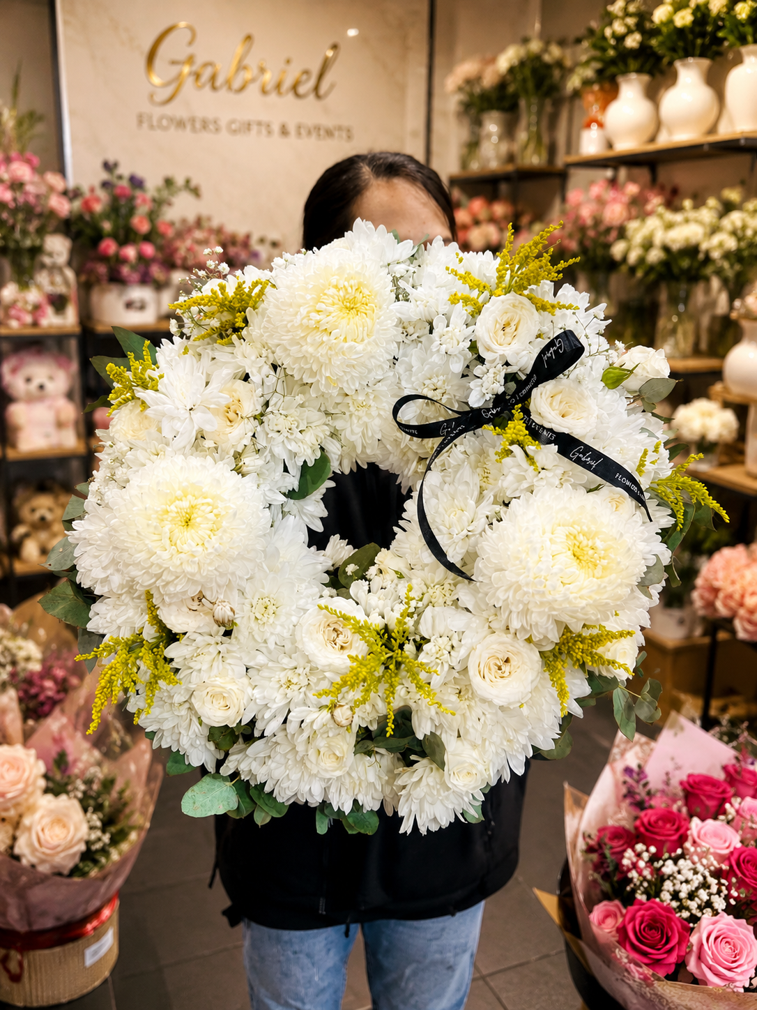 Elegant white funeral wreath handcrafted with premium white chrysanthemums, white roses, seasonal white flowers, golden solidago, and fresh eucalyptus foliage. Designed in a classic open-circle shape to symbolise eternal love and remembrance, this sympathy wreath is finished with a black satin ribbon and displayed in a modern flower shop. A timeless floral tribute suitable for funeral services, memorials, celebrations of life, and heartfelt condolences.