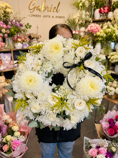 Elegant white funeral wreath handcrafted with premium white chrysanthemums, white roses, seasonal white flowers, golden solidago, and fresh eucalyptus foliage. Designed in a classic open-circle shape to symbolise eternal love and remembrance, this sympathy wreath is finished with a black satin ribbon and displayed in a modern flower shop. A timeless floral tribute suitable for funeral services, memorials, celebrations of life, and heartfelt condolences.
