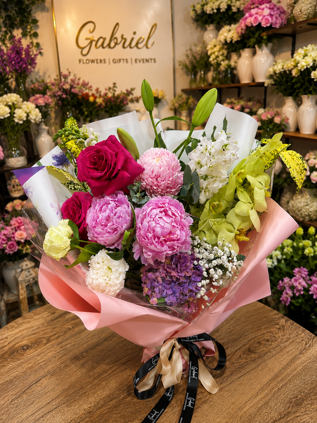 A luxurious hand-tied bouquet featuring vibrant pink roses, lush pink peonies, green cymbidium orchids, white lilies, purple hydrangea, chrysanthemums, stock flowers, and seasonal blooms, beautifully wrapped in premium pink paper with elegant ribbon. A colourful and premium floral arrangement perfect for birthdays, celebrations, anniversaries, Mother's Day, and heartfelt gifting. 🌸💐