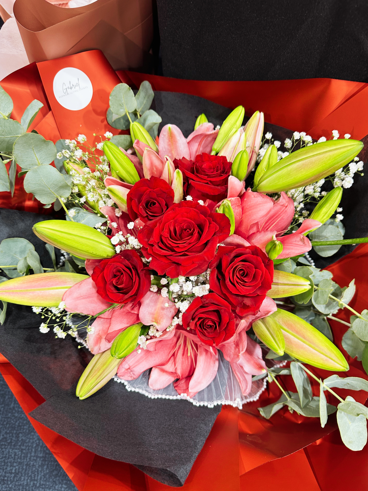 A luxurious round bouquet featuring fresh red roses, fragrant white oriental lilies, and premium seasonal flowers, elegantly wrapped in red and black paper. The romantic floral arrangement combines rich red blooms with soft white lilies, creating a sophisticated and timeless bouquet perfect for anniversaries, Valentine's Day, birthdays, and special celebrations.background