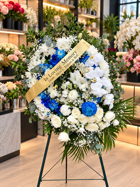 Blue Remembrance premium funeral wreath featuring vibrant blue chrysanthemums, elegant white Phalaenopsis orchids, ivory roses, white chrysanthemums, delicate baby's breath and lush eucalyptus greenery, finished with a personalised memorial sash and displayed on a standing easel against a softly blurred flower shop background.