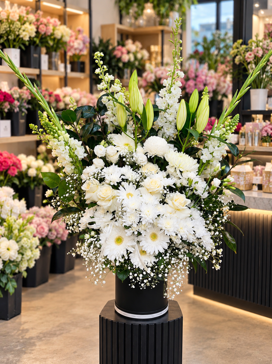 Eternal Grace luxury flower box featuring ivory-white roses, white chrysanthemums, gerberas, Oriental lily buds, white stock flowers, baby's breath and lush greenery, elegantly arranged in a premium black round flower box against a softly blurred florist shop background.