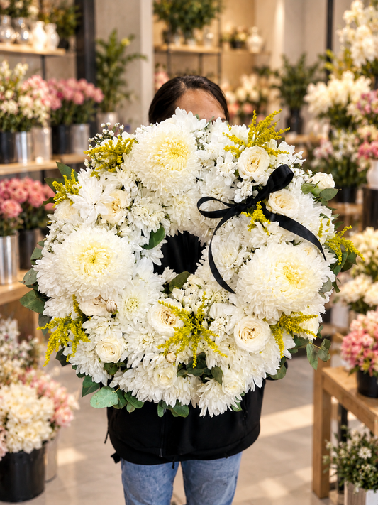 Eternal Peace white chrysanthemum and rose funeral wreath featuring large white chrysanthemums, ivory roses, delicate white blooms, golden-yellow accents, eucalyptus greenery and a black ribbon, beautifully presented in an elegant flower shop.