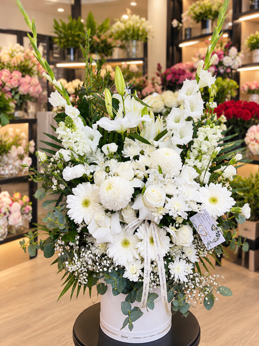 Heavenly Elegance luxury flower box featuring white lilies, ivory roses, chrysanthemums, white gerberas, tall gladiolus, delicate baby's breath and lush eucalyptus greenery, beautifully arranged in an elegant white round flower box against a softly blurred flower shop background.