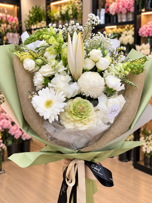 Ivory Bliss premium white lily and rose bouquet featuring ivory roses, Oriental lily buds, white chrysanthemums, gerberas, snapdragons, ornamental kale, baby's breath and lush greenery, beautifully wrapped in sage-green paper with elegant ribbons against a softly blurred flower shop background.