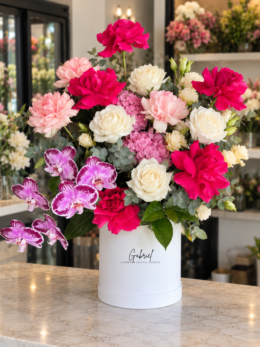 Vibrant Romance premium flower box featuring vibrant hot pink and white roses, blush pink carnations, pink hydrangeas, cascading purple-speckled orchids and fresh eucalyptus greenery, beautifully arranged in an elegant white cylindrical flower box.
